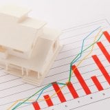 不動産市場の動向分析を示す、白い住宅模型と上昇傾向の棒グラフ、複数折れ線グラフが描かれた資料。