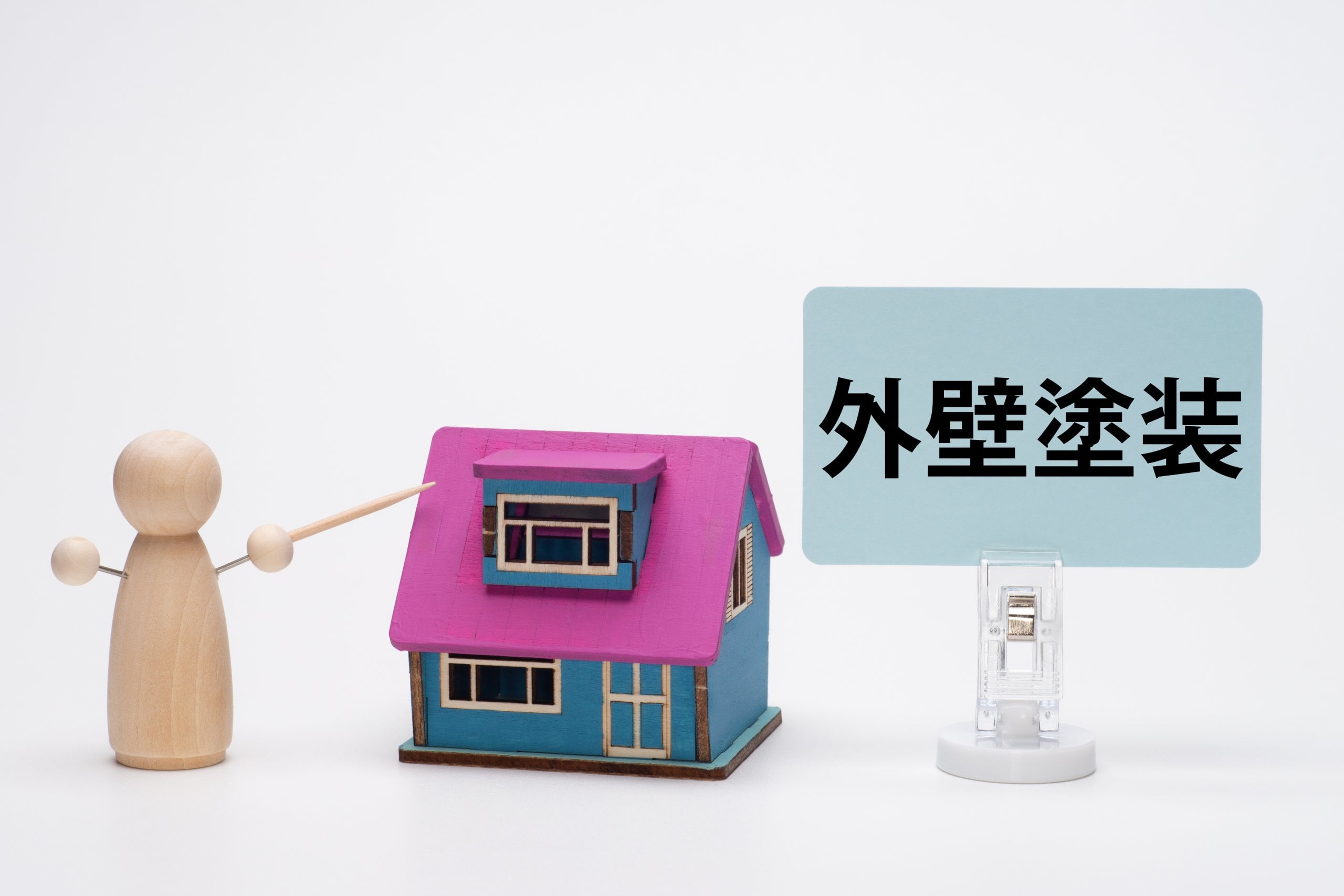 「外壁塗装」と書かれたボードを指し示す木製の人形と、その横に置かれた小さな家の模型